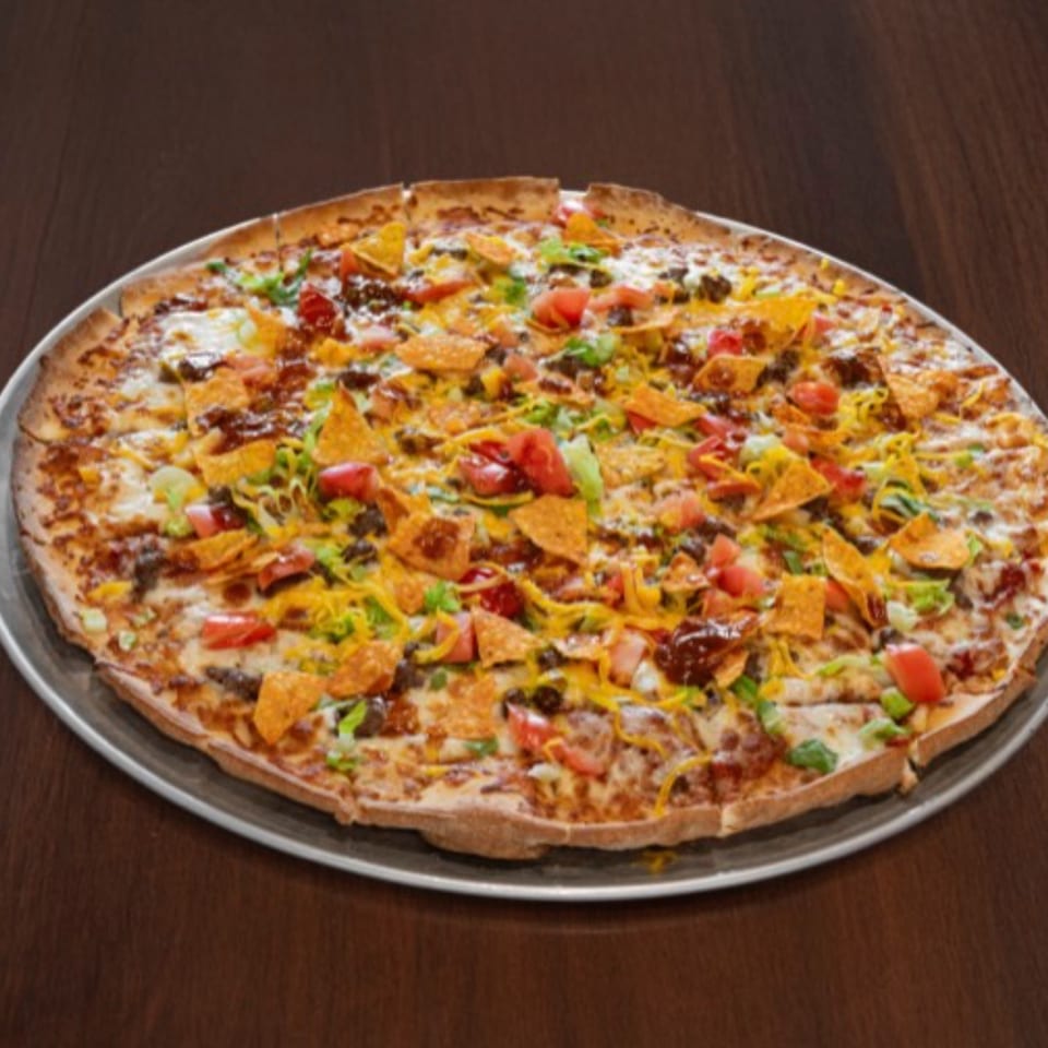 A large taco pizza on a metal pan, topped with cheese, ground beef, tomatoes, and tortilla chips.