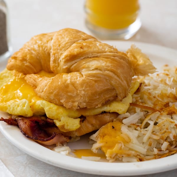 Croissant breakfast sandwich with scrambled eggs, cheese, bacon, and hash browns on a white plate.