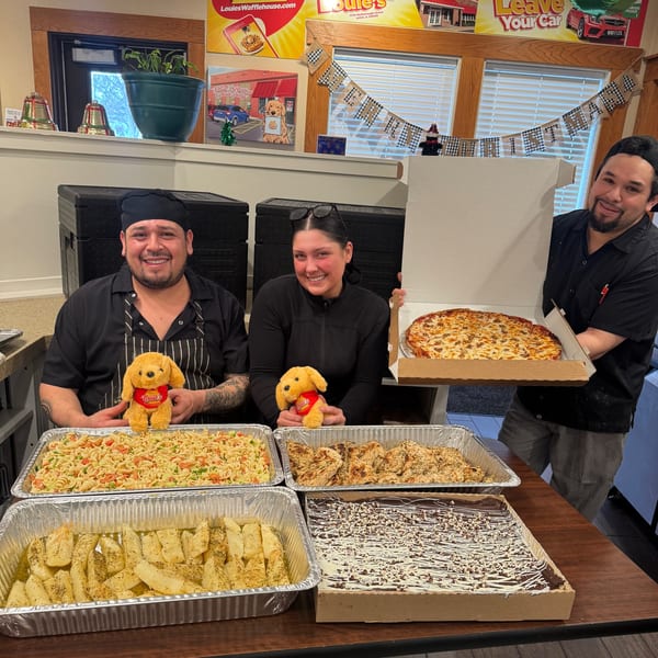 Three Louie's Waffle House staff members smiling with catering trays of pasta, chicken, potatoes, dessert, and a pizza.