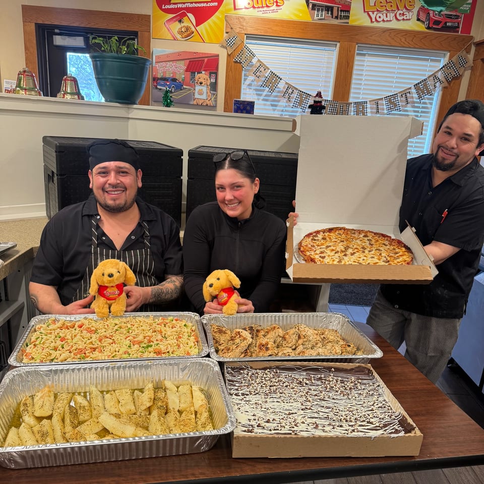 Three Louie's Waffle House staff members smiling with catering trays of pasta, chicken, potatoes, dessert, and a pizza.