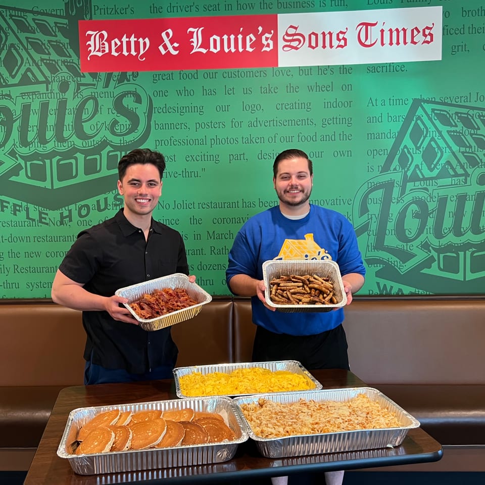 Two men hold large trays of breakfast food, including bacon and French toast sticks, with more food on a table.