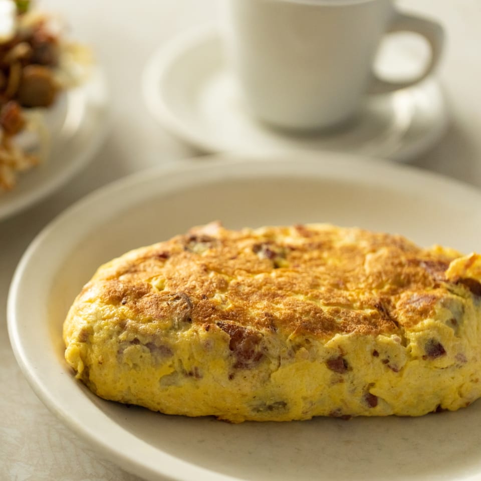 A golden-brown omelet with visible fillings on a white plate, with a coffee cup in the background.