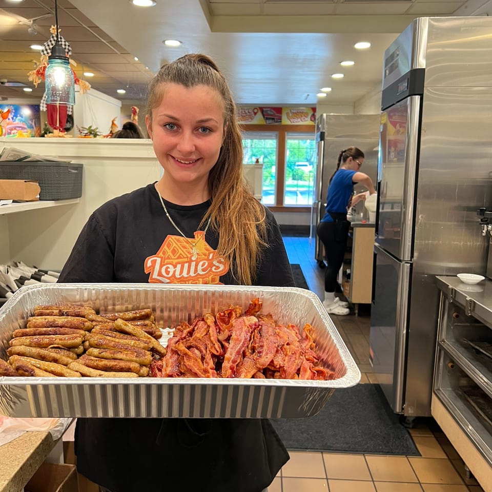 A Louie's Waffle House employee holds a large tray of cooked sausage links and bacon.