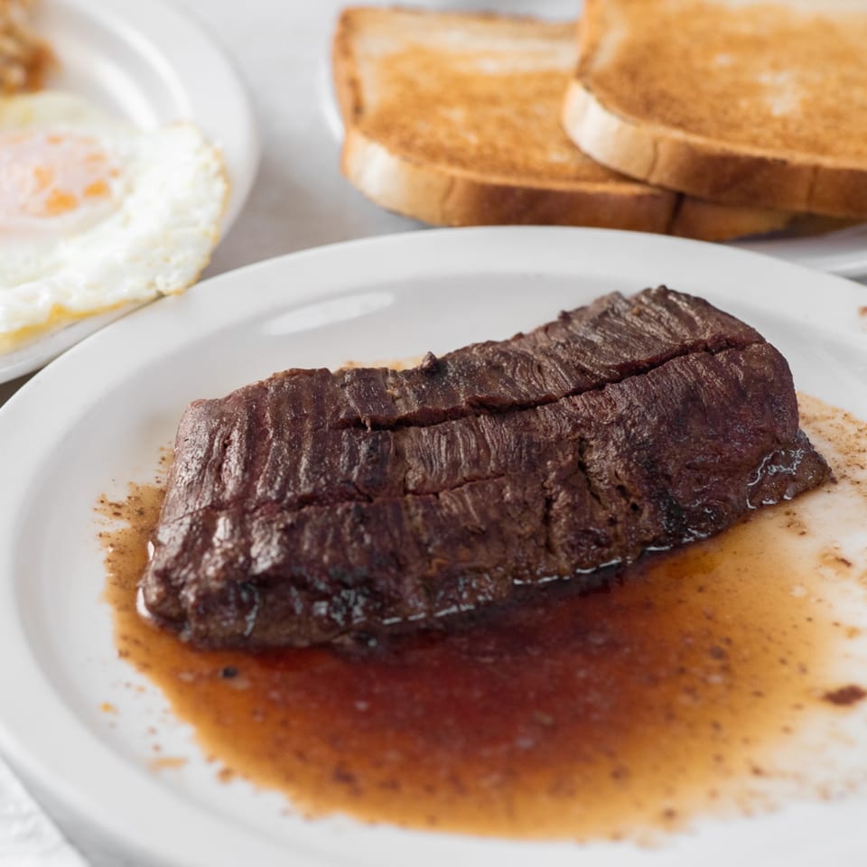 A juicy steak with gravy on a white plate, with fried eggs and toast in the background.