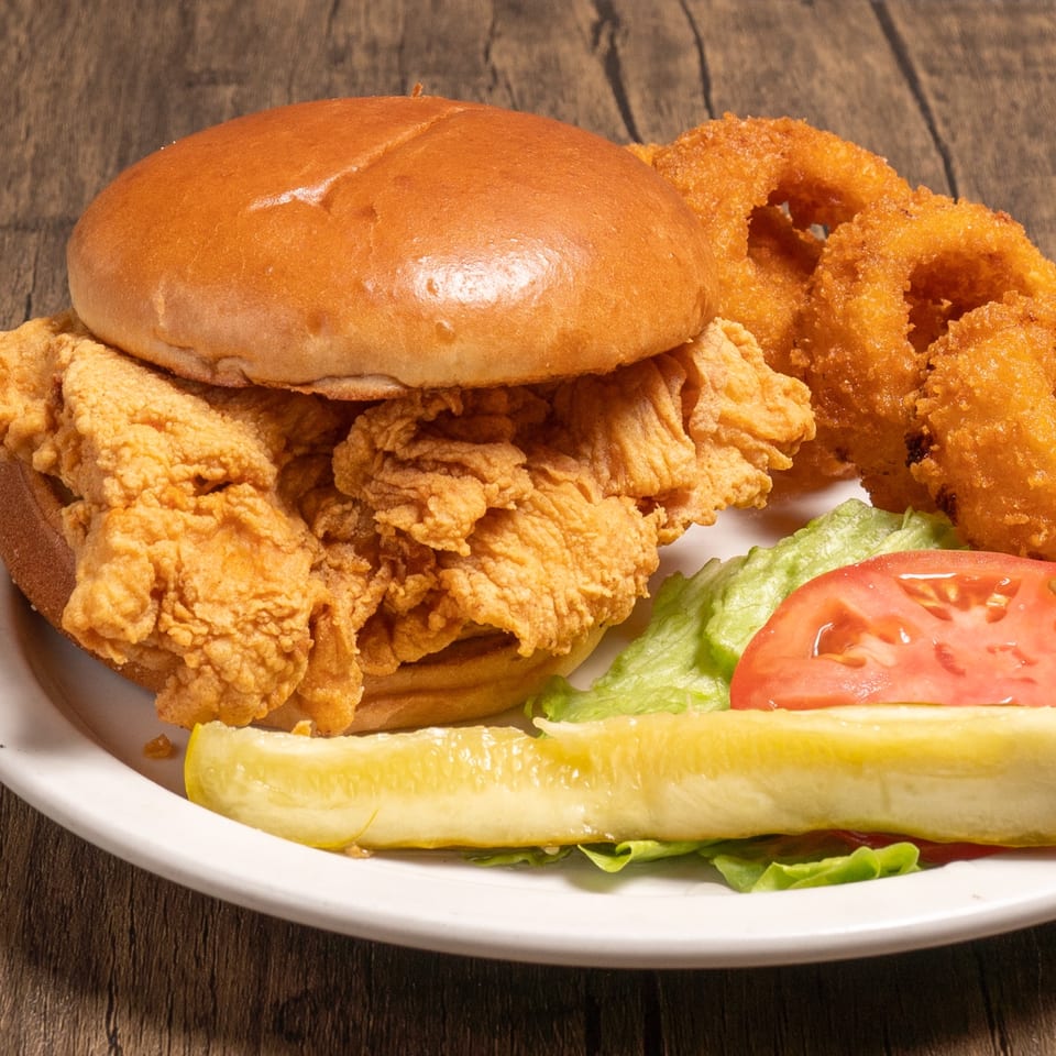 Crispy fried chicken sandwich with onion rings, lettuce, tomato, and a pickle spear.