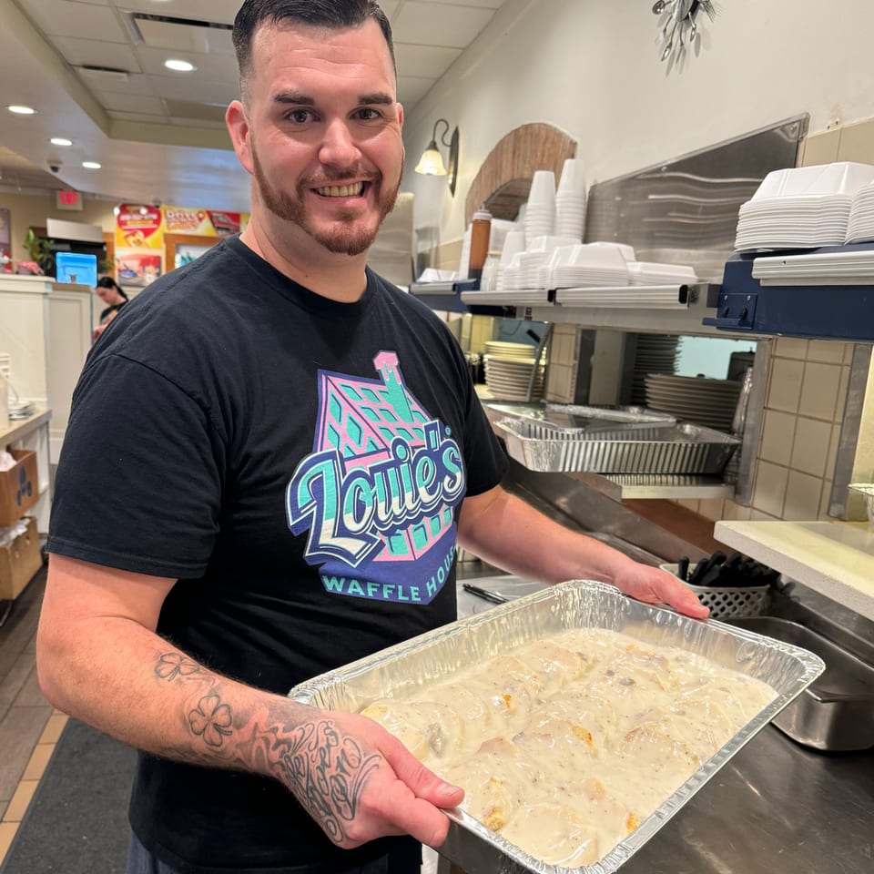 A smiling Louie's Waffle House employee holds a large tray of biscuits and gravy in a kitchen.