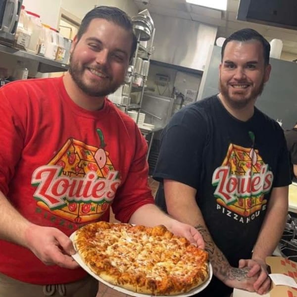 Two smiling men in Louie's Pizza House shirts hold a freshly baked cheese pizza in a kitchen.