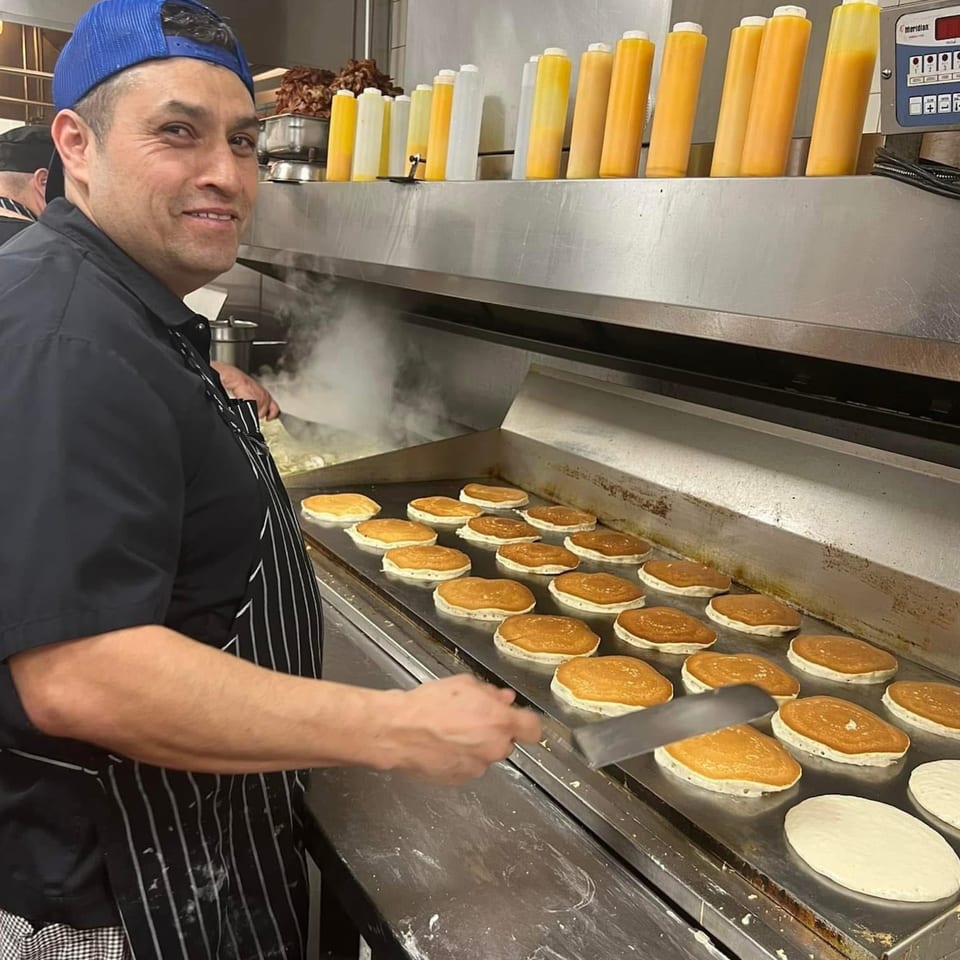 A chef in a blue cap smiles while cooking many pancakes on a griddle at Louie's Waffle House.