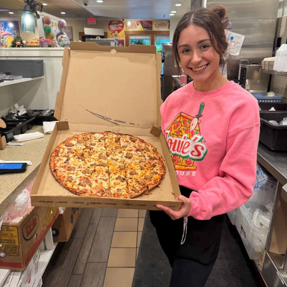 A smiling Louie's Waffle House employee holds a large pizza with sausage in an open box.