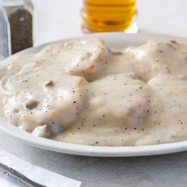 A white plate of biscuits and gravy, with a pepper shaker and drink in the background.