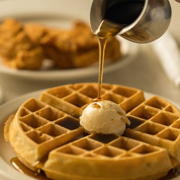 Syrup pouring from a metal pitcher onto a golden waffle with a scoop of butter.