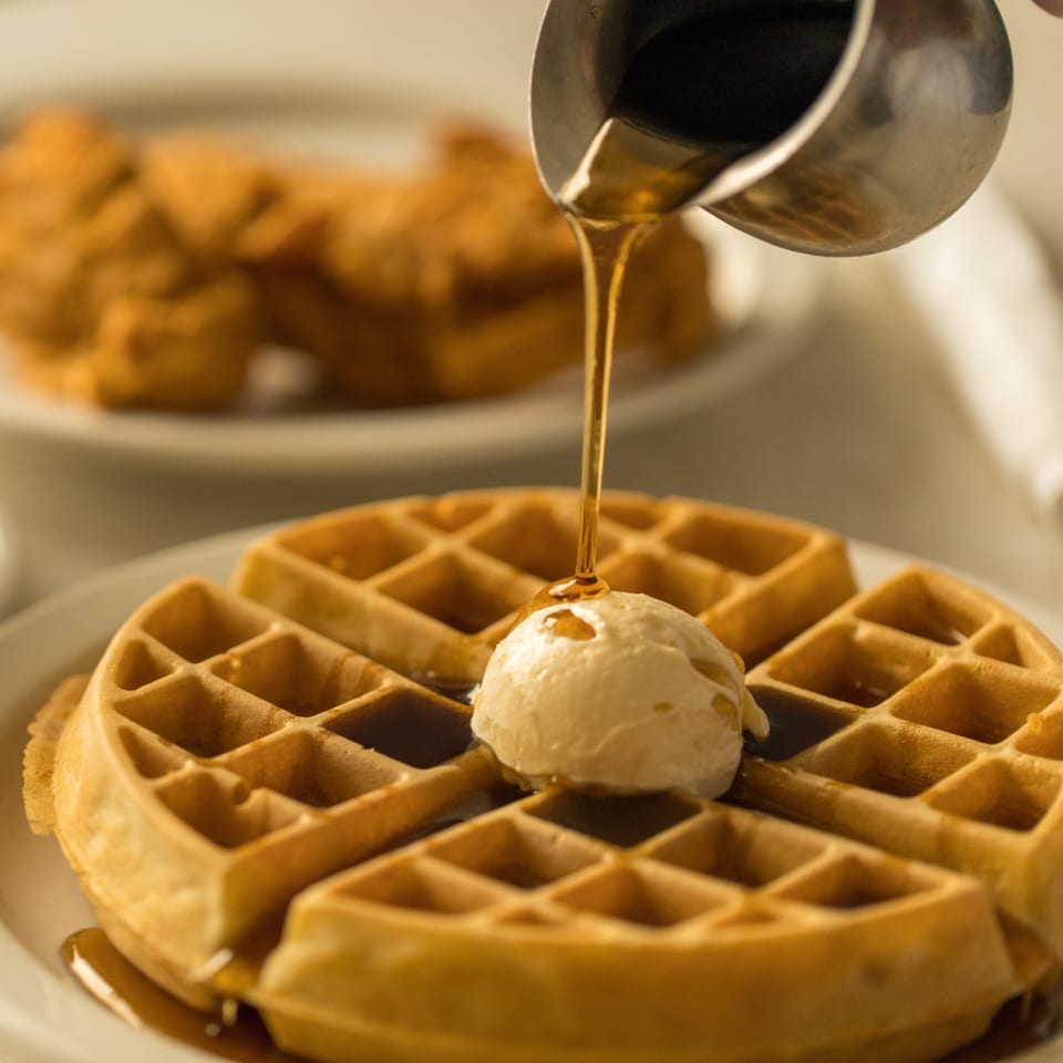 Syrup pouring from a metal pitcher onto a golden waffle with a scoop of butter.