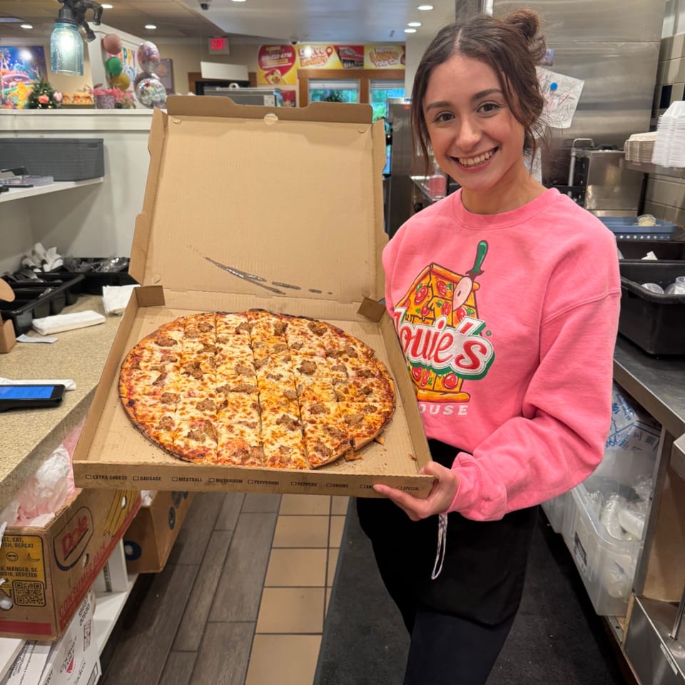 Smiling Louie's Waffle House employee in a pink sweatshirt holding a large sausage pizza.