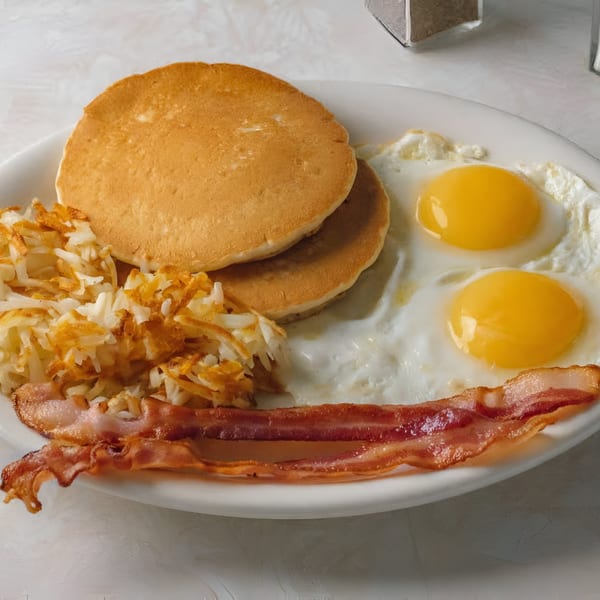 A breakfast plate with pancakes, hash browns, two fried eggs, and crispy bacon.