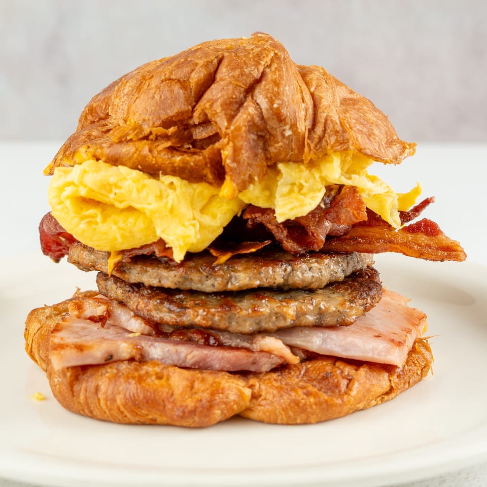 A croissant breakfast sandwich with scrambled eggs, bacon, sausage patties, and ham on a white plate.