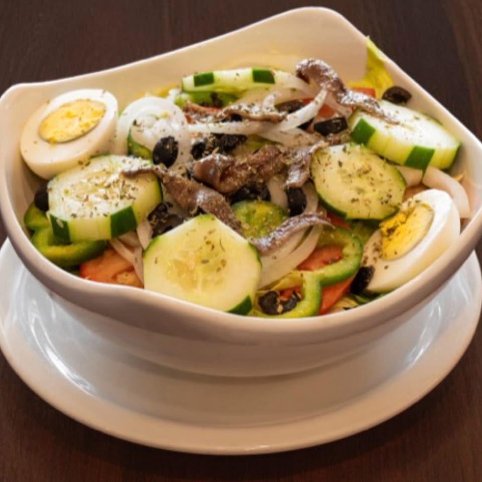 A salad with anchovies, hard-boiled eggs, cucumbers, onions, and bell peppers in a white bowl.