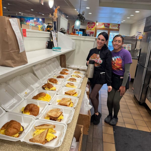 Two Louie's Waffle House employees stand proudly beside a counter filled with breakfast takeout orders.
