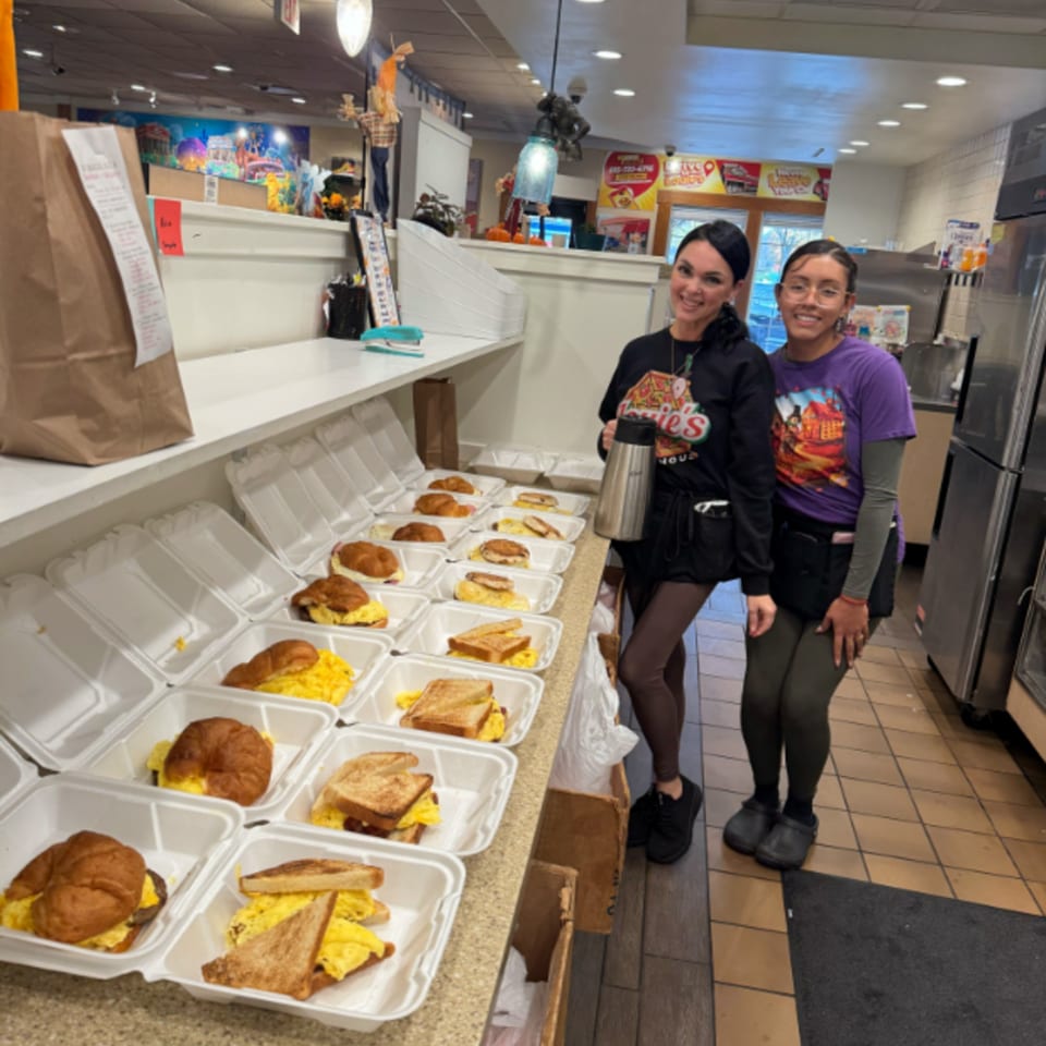 Two Louie's Waffle House employees stand proudly beside a counter filled with breakfast takeout orders.