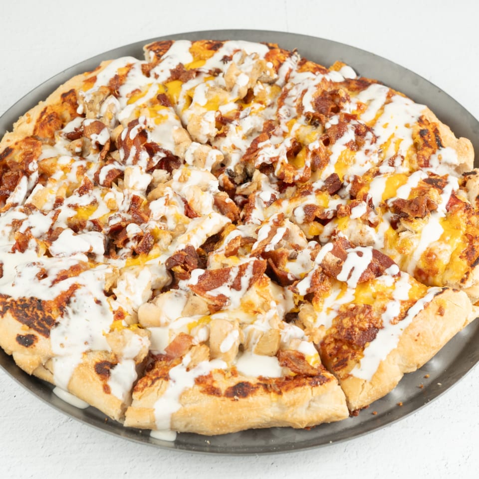 A breakfast pizza with chicken, bacon, cheese, and a white sauce drizzle on a dark metal pan.