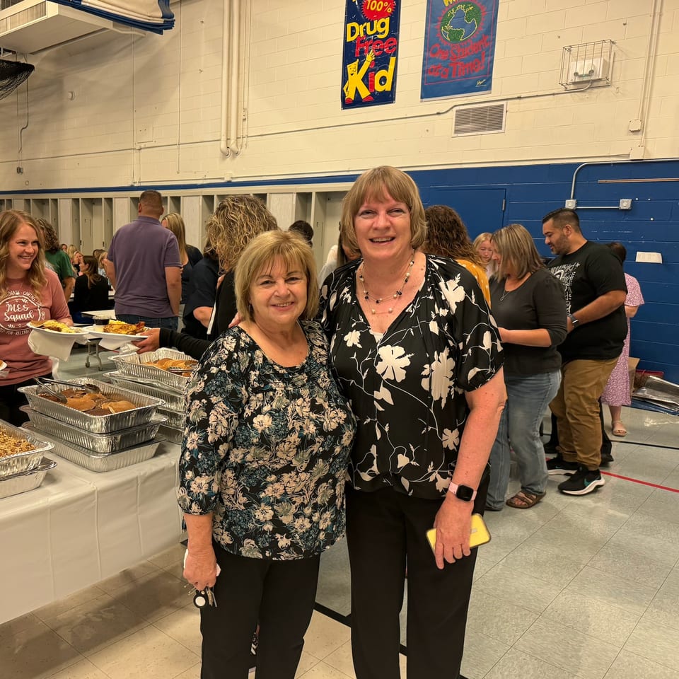 Two women smiling at the camera in front of a buffet line with people in the background.