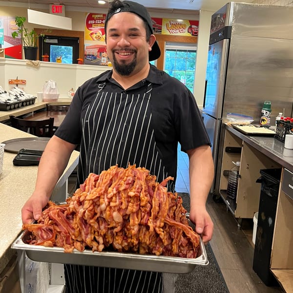 A smiling Louie's Waffle House chef holds a large tray overflowing with crispy bacon in a kitchen.