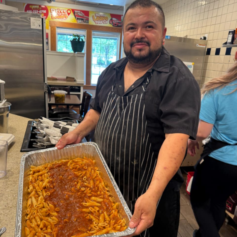 A smiling chef in a black shirt and striped apron holds a large tray of penne pasta with red sauce.