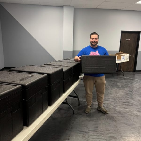 A man in a blue shirt holds a black insulated food carrier next to a table with more carriers.